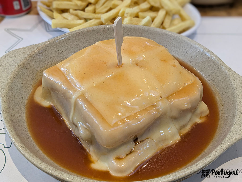 Um prato de francesinha, um sanduíche português coberto com queijo derretido e um rico molho marrom, é servido em uma tigela rasa com um palito de dente em cima. As batatas fritas são visíveis ao fundo - perfeitas para quem está se perguntando onde comer Francesinha no Porto.