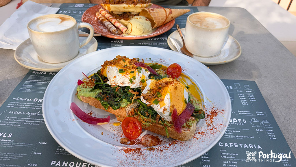 Uma mesa de café preparada para o brunch no Porto, com dois cappuccinos, torrada de abacate com ovos escalfados, verduras e tomates, além de um prato de waffles com um cone de waffle e sorvete ao fundo.