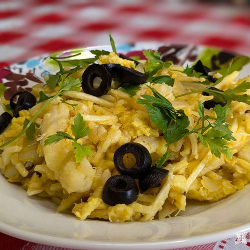Um prato colorido de Bacalhau à Brás Português, uma receita deliciosa, é mostrado com bacalhau salgado desfiado, cebolas, batatas, ovos e guarnecido com azeitonas pretas e salsa fresca. O prato é disposto em um prato decorativo, sobre uma toalha de mesa xadrez vermelha e branca.