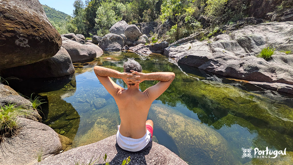 uma pessoa observa um lago esverdeado muito sereno rodeado por pedras