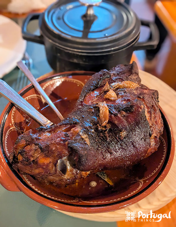 A ceramic dish holds a roasted pork leg with dark, crispy skin, garnished with garlic and herbs. A metal spoon and fork sit inside the plate, a reminder of what you might enjoy while exploring the culinary delights of Seia. In the background, a black pot rests on the table.