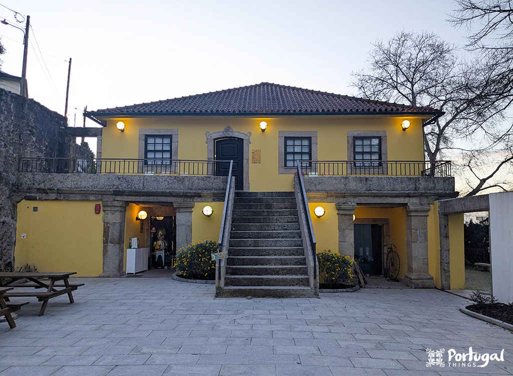 A small yellow building with a tiled roof and central staircase, surrounded by trees, offers a charming stop if you are wondering what to do in Seia. Its symmetrical design features double doors and windows. Outdoor lights glow warmly on the paved area with a picnic table out front.