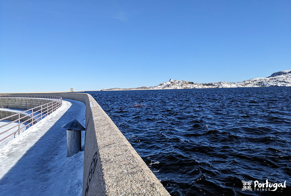 Snow covers the walkway next to a large body of dark blue water, surrounded by a stone wall. A vast, clear blue sky and a snow-covered landscape stretch out in the background.