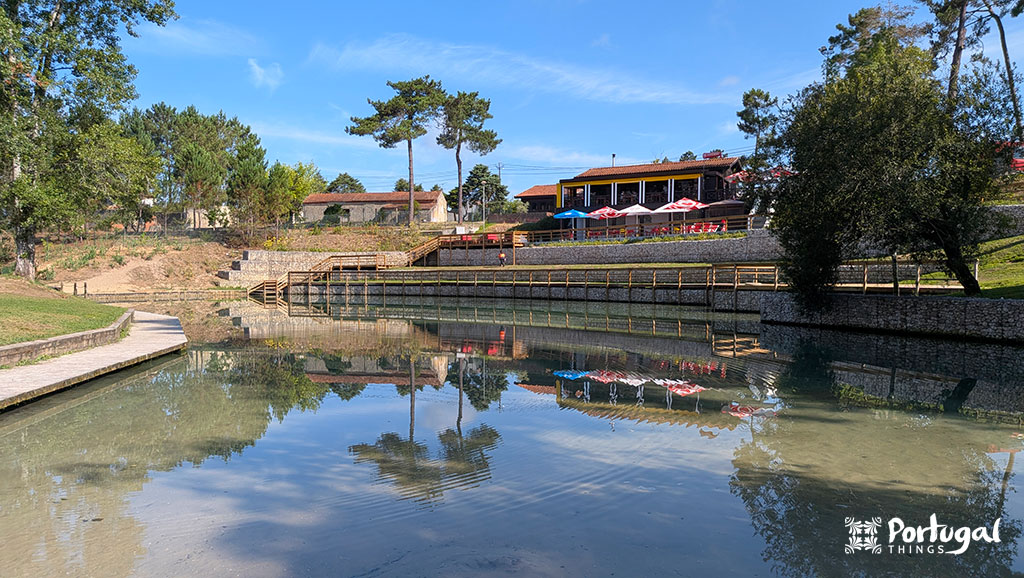 Olhos de Fervença river beach with blue-green water, a café in the background, and seating areas on the grass.