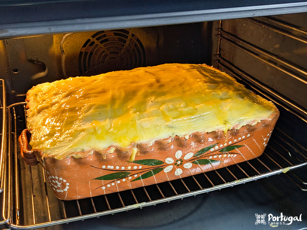 A rectangular clay dish with a decorative pattern contains a large casserole of Bacalhau Escondido covered in egg yolk, baking in the oven.