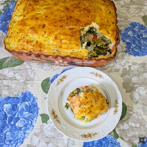 Uma caçarola retangular assada, Bacalhau Escondido, com uma crosta dourada, está sobre uma toalha de mesa floral. Uma porção foi cortada e colocada em um prato branco, revelando camadas de vegetais dentro dessa deliciosa receita de bacalhau.