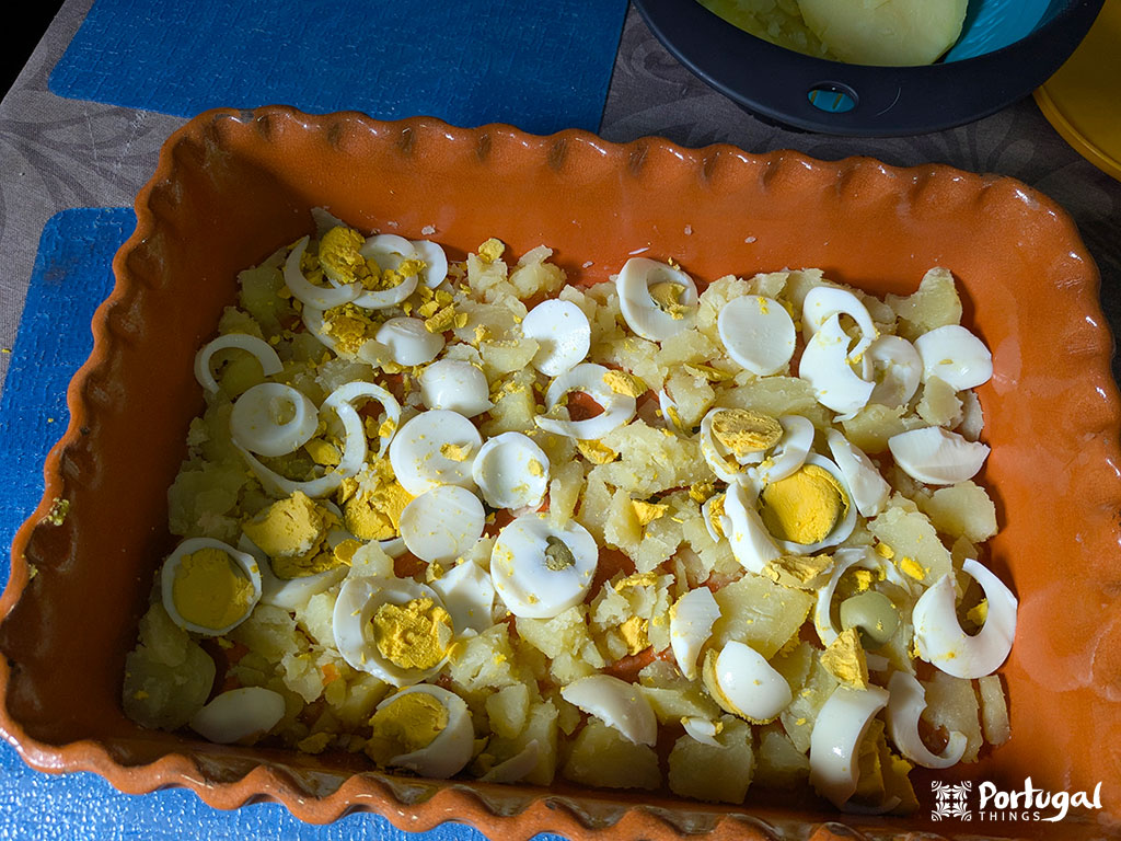 An orange baking dish filled with sliced ​​boiled eggs and chopped boiled potatoes, arranged in a single layer, resembling a Bacalhau Escondido.