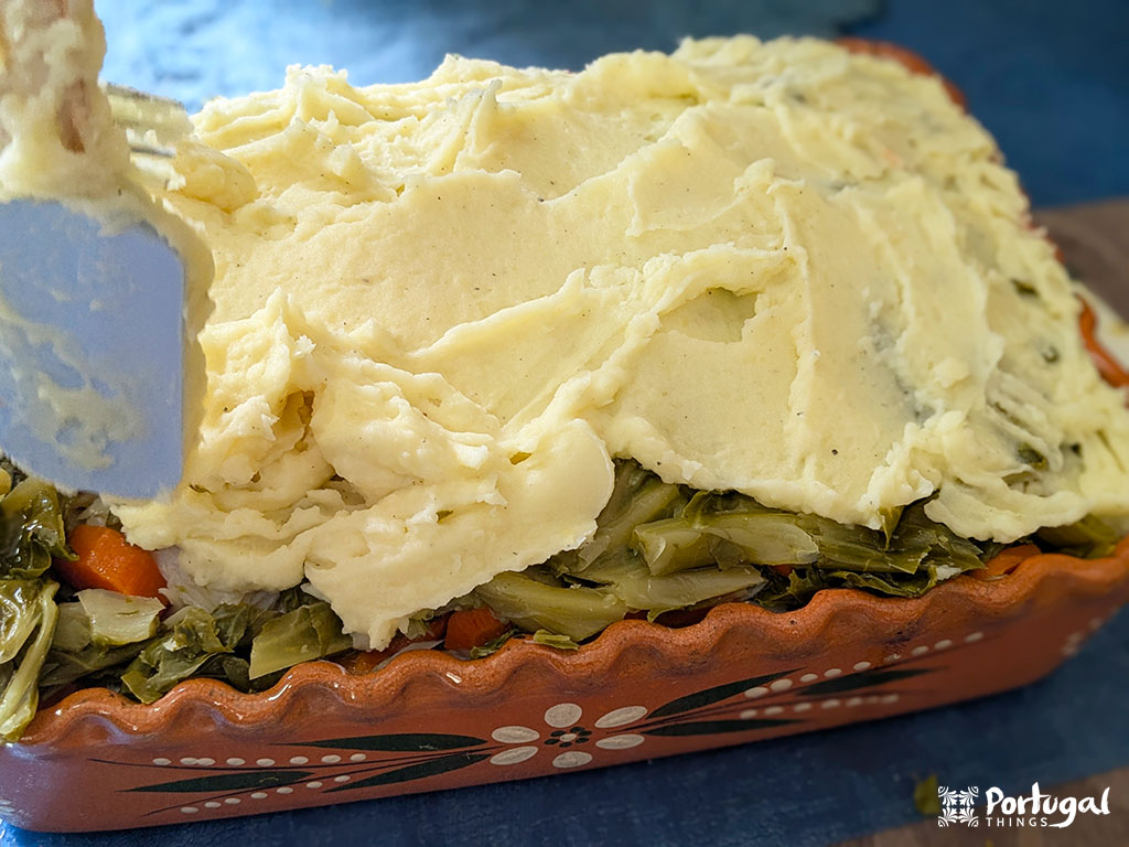 A clay dish with cooked vegetables is being covered with mashed potatoes using a spatula, creating a hidden cod recipe.