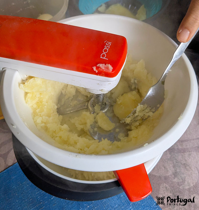 A red-handled potato masher is used to mash boiled potatoes in a white bowl, perfect for preparing the Cod Recipe. A fork rests inside the bowl as steam escapes from the potatoes.