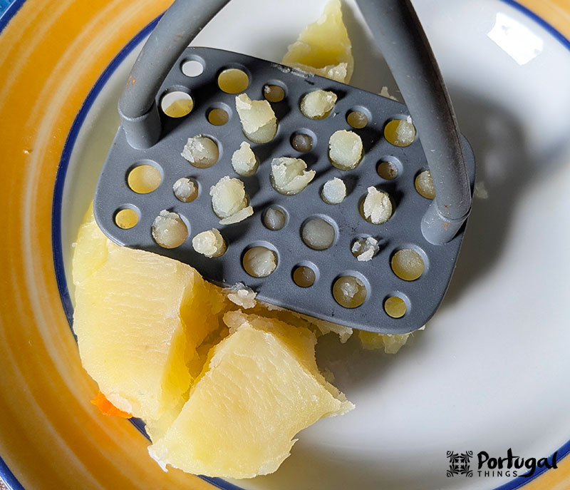 A metal potato masher is pressing cooked potatoes onto a white plate with a yellow and blue border. Perfect for preparing Bacalhau Escondido, the potatoes are partially mashed