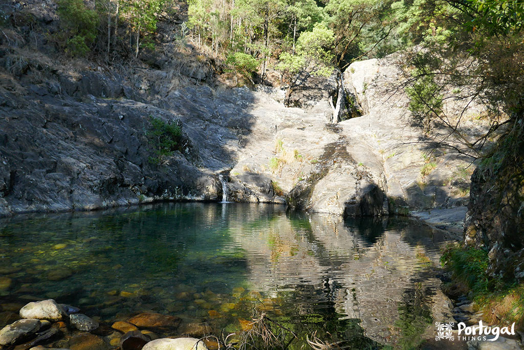 A small waterfall cascades down rocky cliffs into a clear, shallow pool surrounded by rocks and green trees, reflecting the landscape—a serene spot among the waterfalls of northern Portugal.
