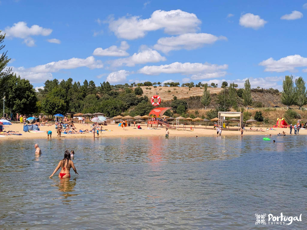 People are swimming and relaxing on a sandy lakeside beach, enjoying one of Portugal's beautiful river beaches. Trees, grassy hills, and playground equipment fill the landscape. The sky is mostly clear, with a few clouds.