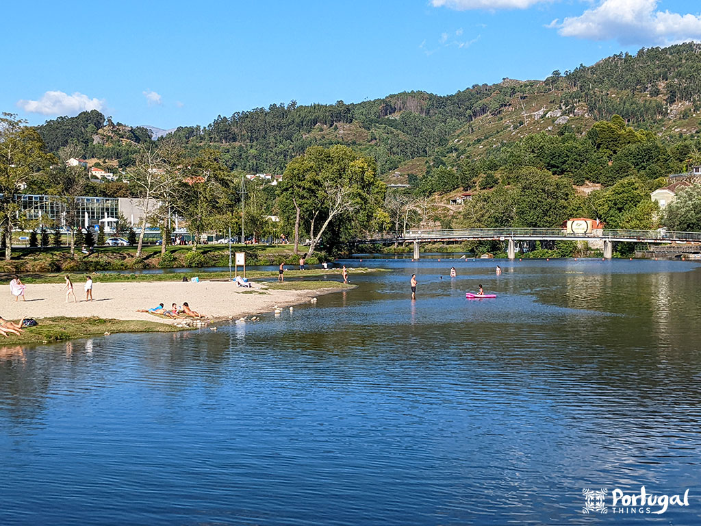 A river with people swimming, kayaking, and standing on a sandy beach—on the river beaches of Valletta, Portugal. Trees, hills, houses, and a bridge appear in the background under a blue sky.