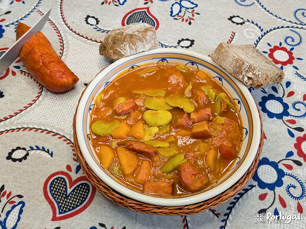Uma tigela de Favas com Chouriço, uma receita tradicional portuguesa, apresenta fatias de chouriço e legumes em uma toalha de mesa estampada. Pão e chouriça inteira ficam ao lado da tigela.