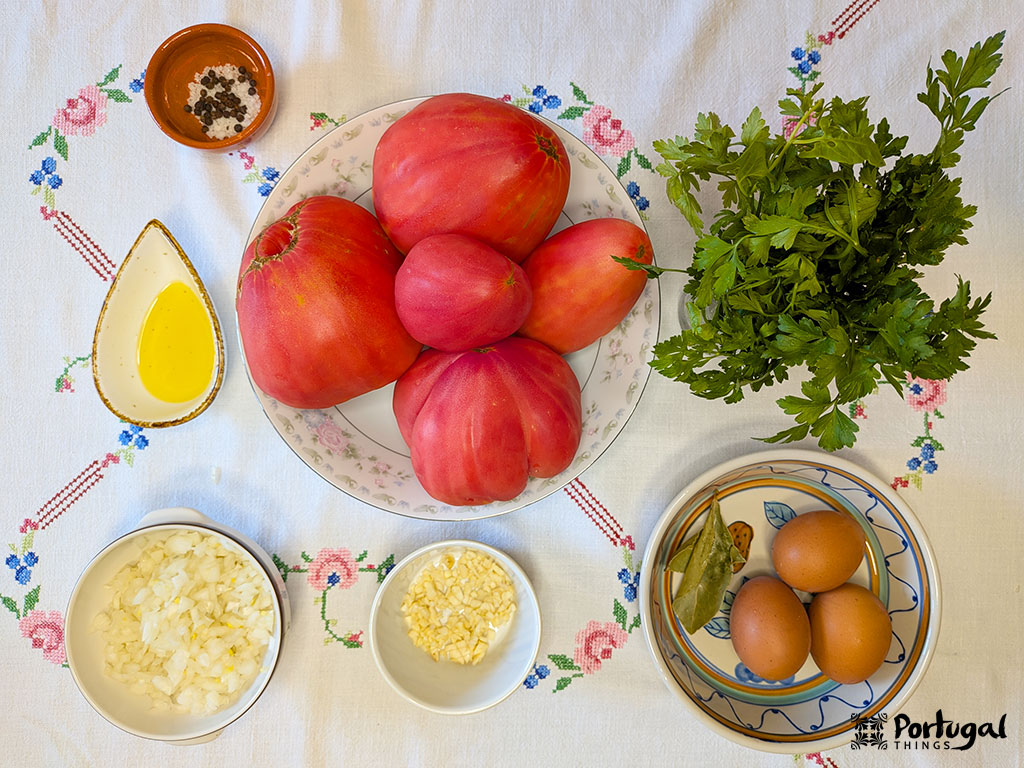 Ingredientes numa mesa para uma Receita Tomatada Alentejana clássica: cinco tomates grandes, salsa, três ovos com folhas de louro, cebola e alho picados, azeite de oliva e um pequeno prato de pimenta preta.