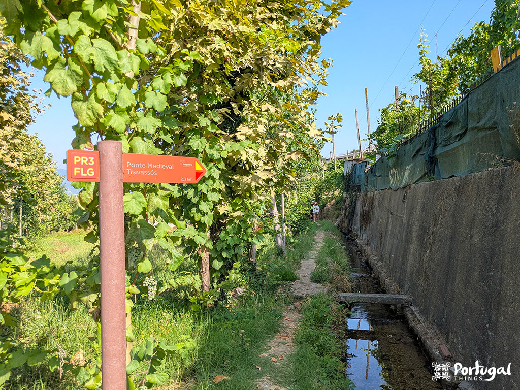 Um estreito caminho de terra passa ao lado de um canal de irrigação de pedra, cercado por vegetação verde. Uma placa de sinalização marca a trilha de caminhada PR3