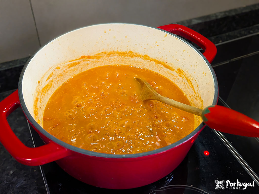 Uma panela vermelha num fogão contém um molho laranja borbulhante que está sendo mexido com uma colher de pau. Inspirado na Receita Tomatada.