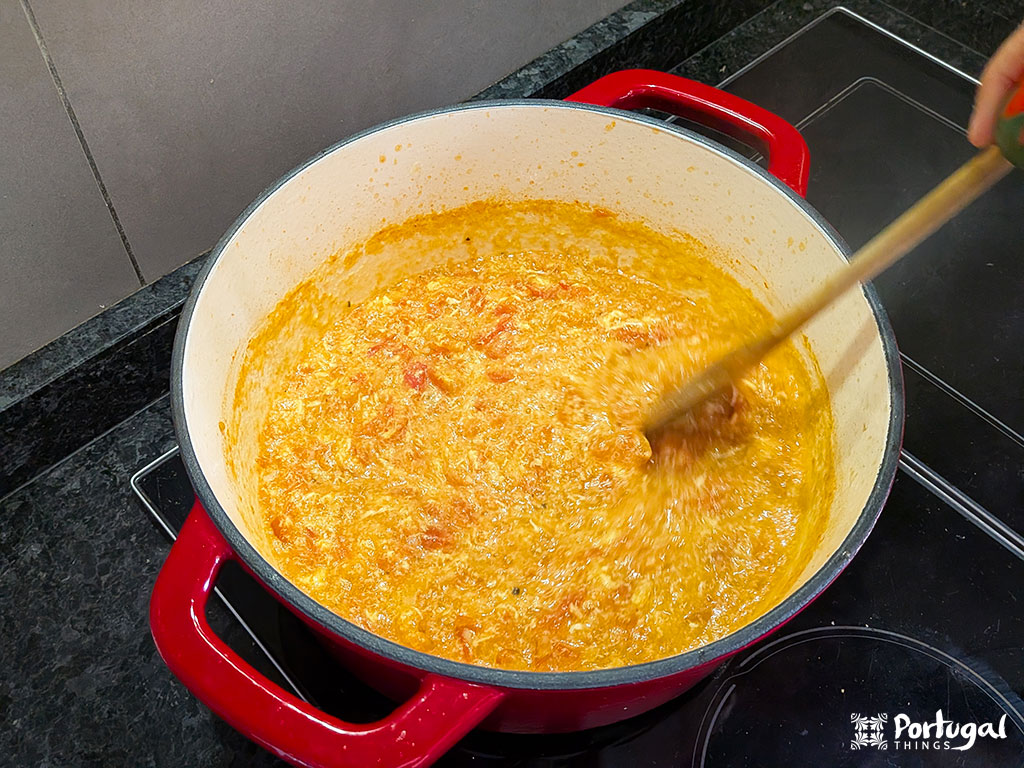Uma panela vermelha em cima de um fogão preto contém uma mistura amarelo-alaranjada que está sendo mexida com uma colher de pau. O ensopado espesso e texturizado lembra a Tomatada à Alentejana.