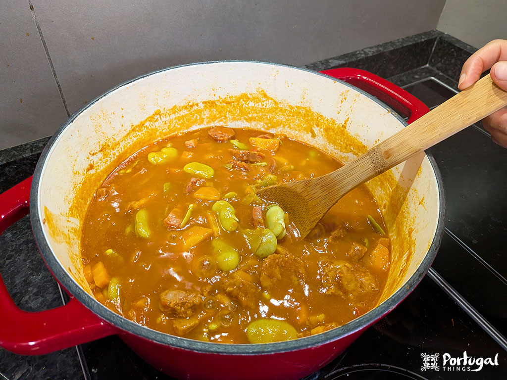 A red pot filled with a thick, orange stew of vegetables, beans, and chunks of meat—Favas com Chouriço, a savory Portuguese recipe—is being stirred with a wooden spoon on a black stovetop.