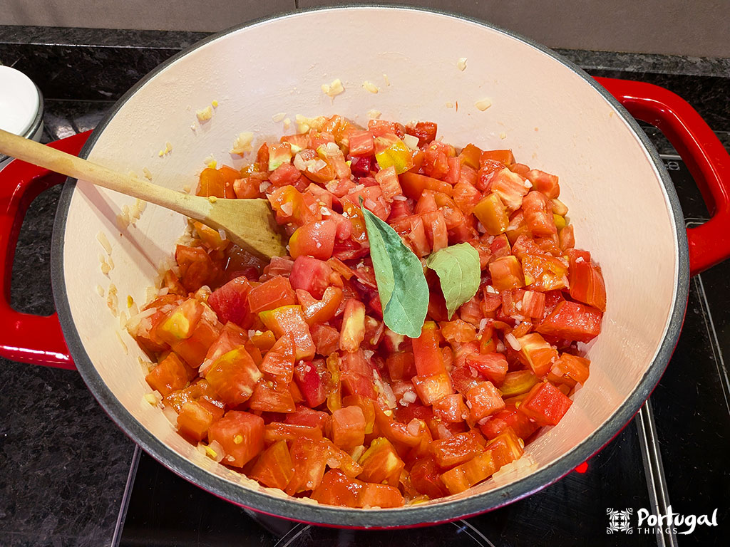 Uma panela vermelha no fogão contém tomates picados, cebolas em cubos, alho picado e folhas de louro, sendo mexidos com uma colher de pau - um estágio inicial da clássica Tomatada à Alentejana.