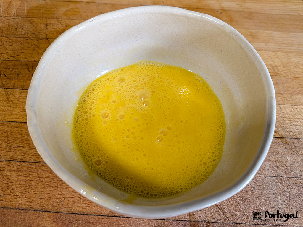 Uma tigela de cerâmica branca contendo ovos batidos com bolhas na superfície, colocada sobre uma bancada de madeira. Ideal para preparar Receita Tomatada à Alentejana.