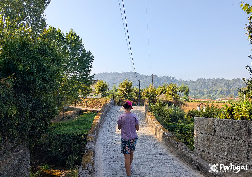 Uma pessoa de camisa rosa caminha pela estreita ponte de pedra na trilha PR3 Levadas de Jugueiros, em Felgueiras, cercada por uma vegetação exuberante e colinas distantes.