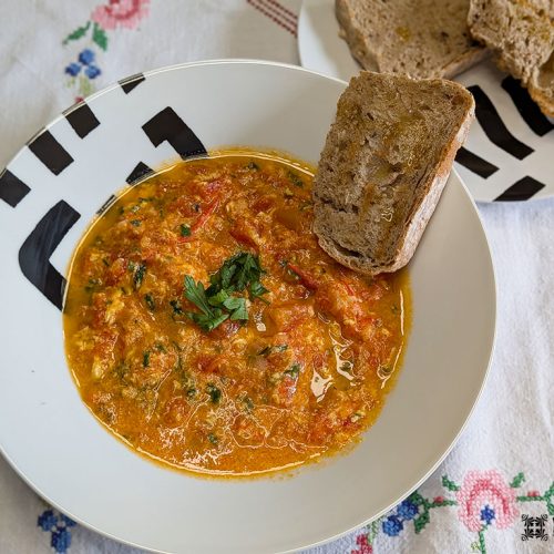 Uma tigela de Receita Tomatada, um prato clássico alentejano à base de tomate, é decorada com ervas aromáticas e servida com pão rústico. Ao fundo, fatias extras repousam sobre uma toalha de mesa branca adornada com bordados florais.