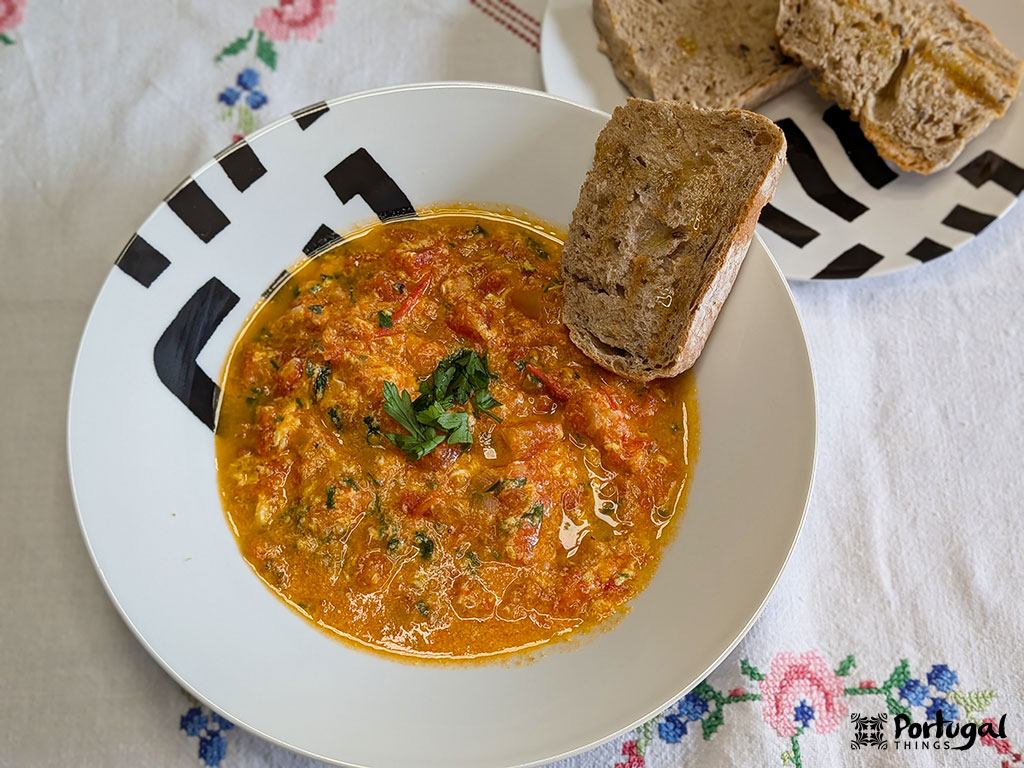 Uma tigela de Receita Tomatada, um prato clássico alentejano à base de tomate, é decorada com ervas aromáticas e servida com pão rústico. Ao fundo, fatias extras repousam sobre uma toalha de mesa branca adornada com bordados florais.