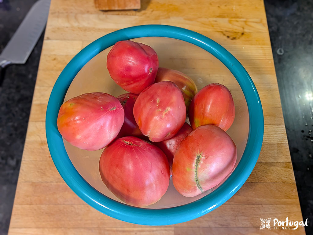 Uma tigela de borda azul cheia de tomates grandes, vermelhos e rosados, perfeitos para uma Receita Tomatada Alentejana, está sobre uma tábua de corte de madeira, com uma faca visível ao fundo.