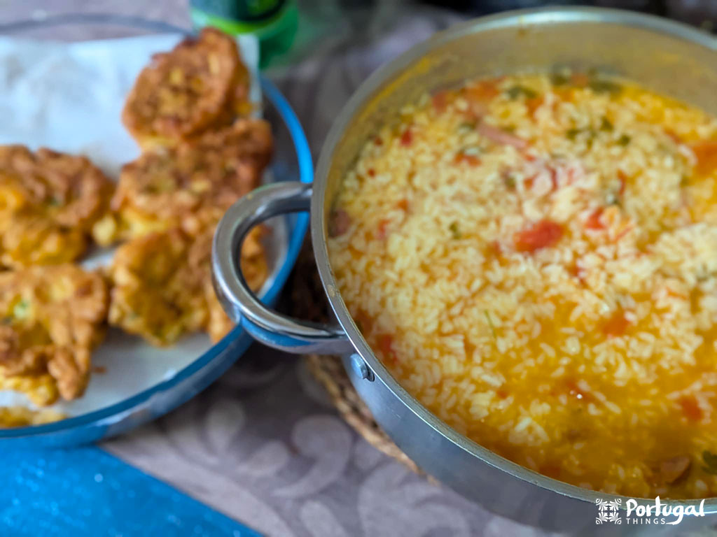 Uma panela de Arroz de Tomate com legumes fica ao lado de um prato de bolinhos fritos dourados em uma mesa coberta por um pano. 