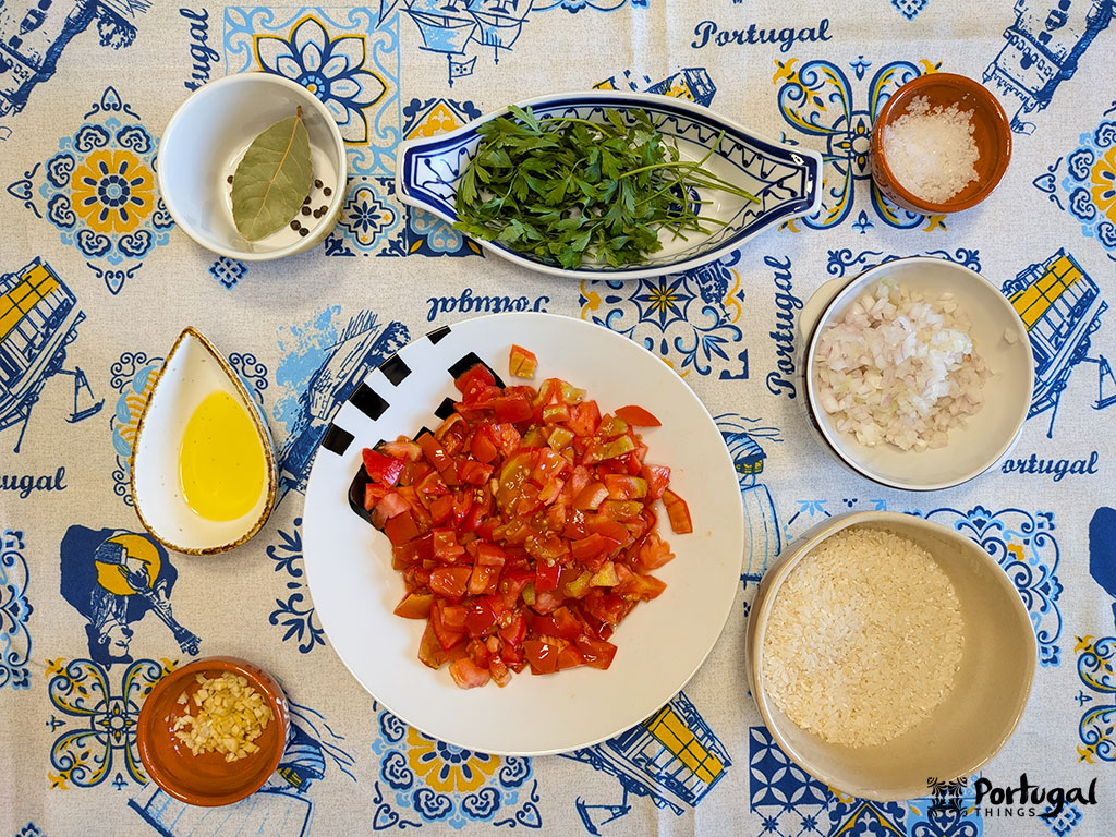 Uma tigela de tomates picados é cercada por pequenos pratos de ingredientes para o Arroz de Tomate - cebola picada, alho picado, azeite de oliva, uma folha de louro com grãos de pimenta, sal marinho, ervas frescas e arroz cru em uma toalha de mesa com tema de Portugal.