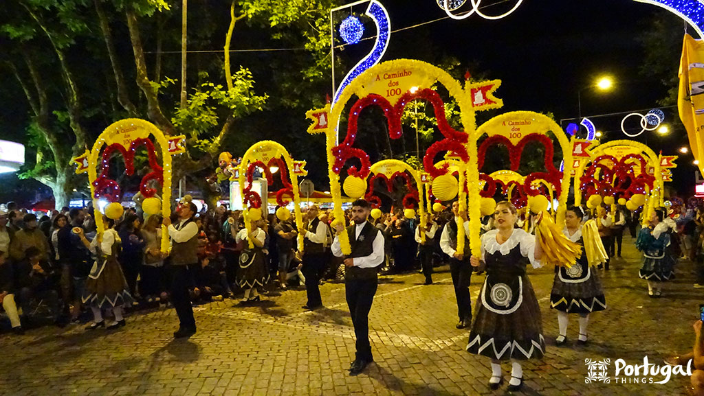 A group of people in traditional attire parades at night, carrying large colorful decorative arches with “100” and “One Hundred Years,” while a crowd watches — one of the many reasons to fall in love with Portugal. Festive lights and trees line the street.