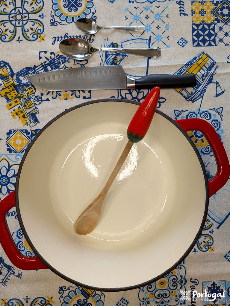 Uma panela esmaltada branca com alças vermelhas fica sobre uma toalha de mesa estampada - perfeita para preparar o Arroz de Tomate. Dentro, há uma colher de pau com cabo vermelho e, acima, utensílios variados, prontos para ajudá-lo a preparar essa receita clássica.