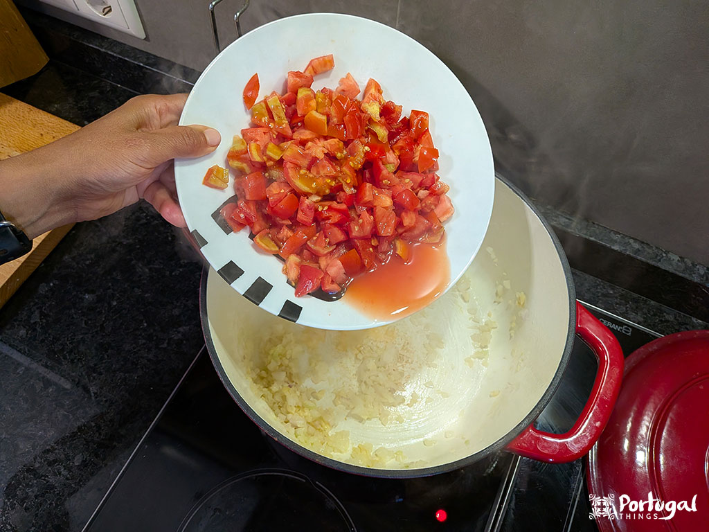 Uma mão segura um prato de tomates picados sobre uma panela com cebolas salteadas, pronta para adicioná-los à receita de Arroz de Tomate. O vapor sobe da panela no fogão, e uma tampa vermelha está próxima.