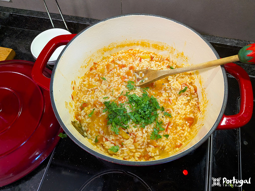 Uma grande panela vermelha  num fogão preto contém Arroz de Tomate misturado coberto com salsa picada. Uma colher de pau está dentro da panela. A tampa da panela está próxima.