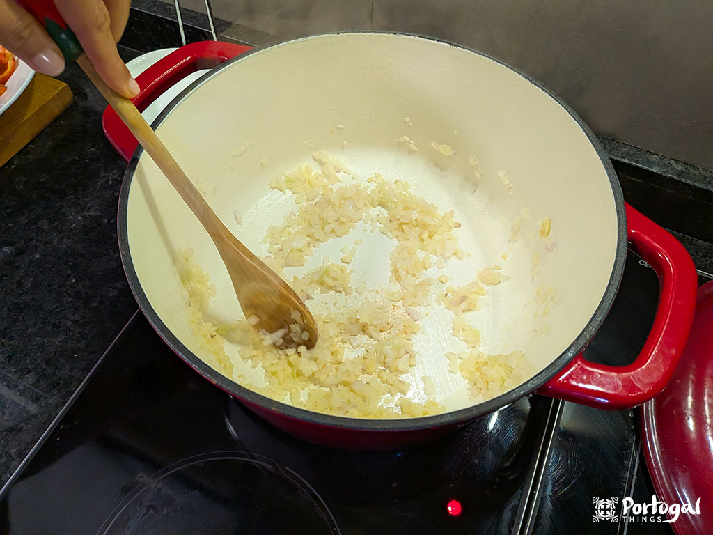 Uma pessoa mexe cebolas picadas numa panela grande de esmalte vermelho num fogão preto, iniciando uma receita de arroz. As cebolas estão sendo salteadas até ficarem translúcidas, com o vapor subindo como o primeiro passo para um Arroz de Tomate ou outra receita fácil.