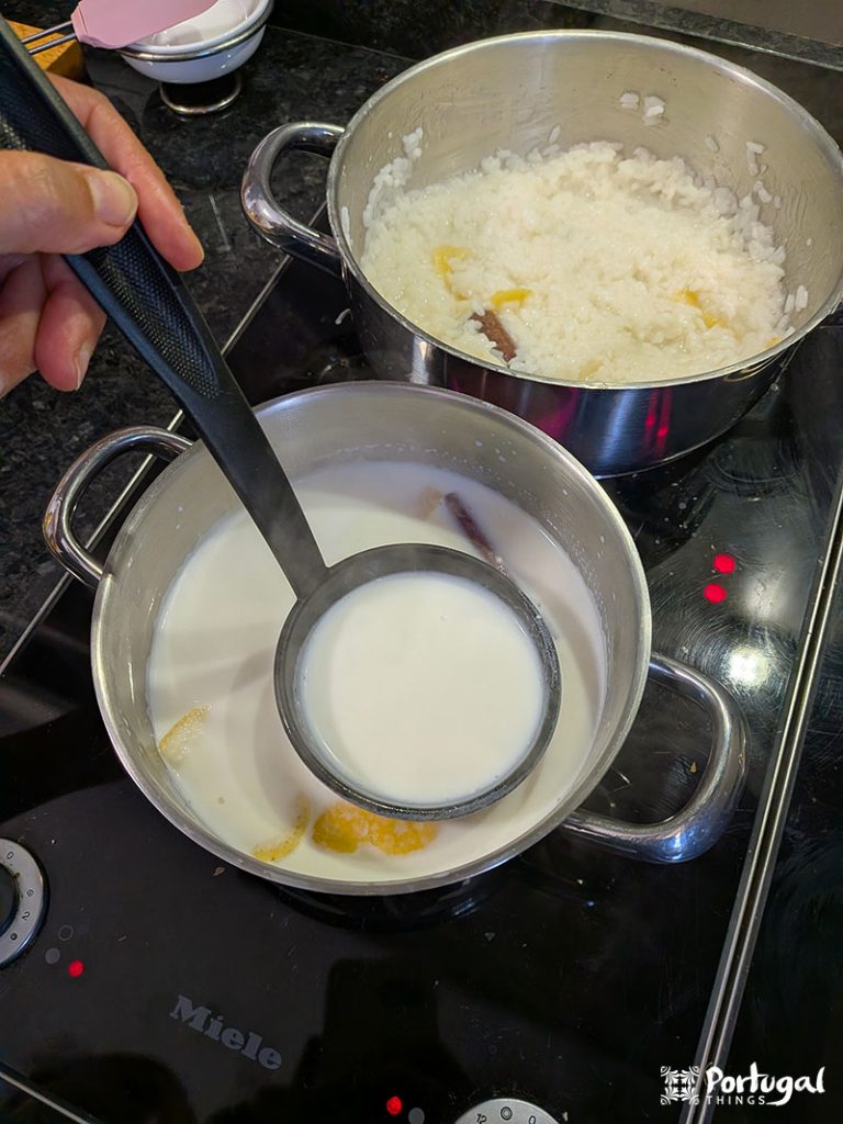 A person holds a ladle over a pot of milk with lemon zest and cinnamon, preparing Arroz-Doce, a traditional recipe. Behind her, a pot of cooked rice sits on an electric stove; both pots sit on a black countertop.