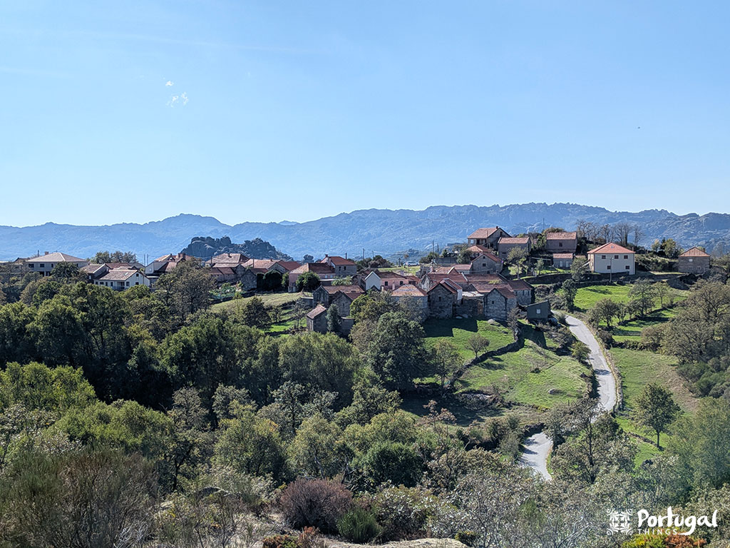 A rural village with red-roofed houses nestles among green hills and trees along the Castrejo Trail. A winding road leads to distant mountains beneath a clear blue sky.