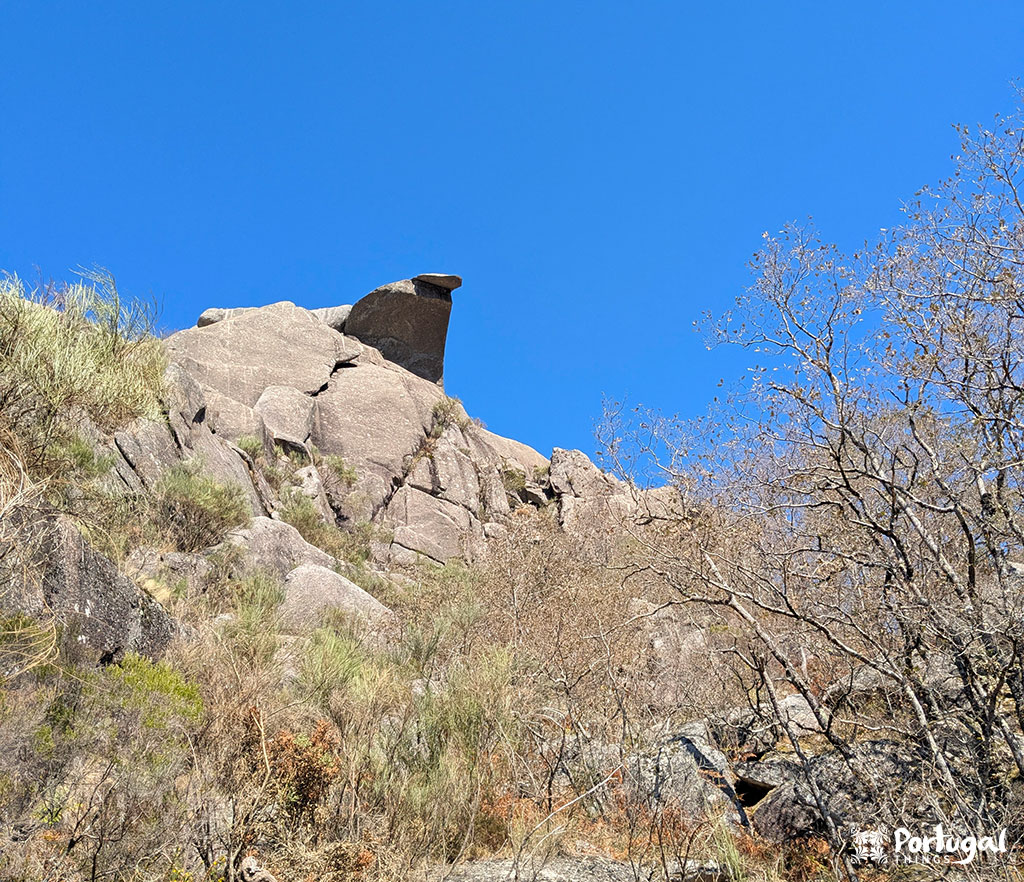 A rock formation with sparse dry vegetation and a clear blue sky. The distinctive rock formation at the top marks the rugged landscape along the PR3 Trilho Castrejo trail in Castro Laboreiro, captured in a dry, sunny climate.