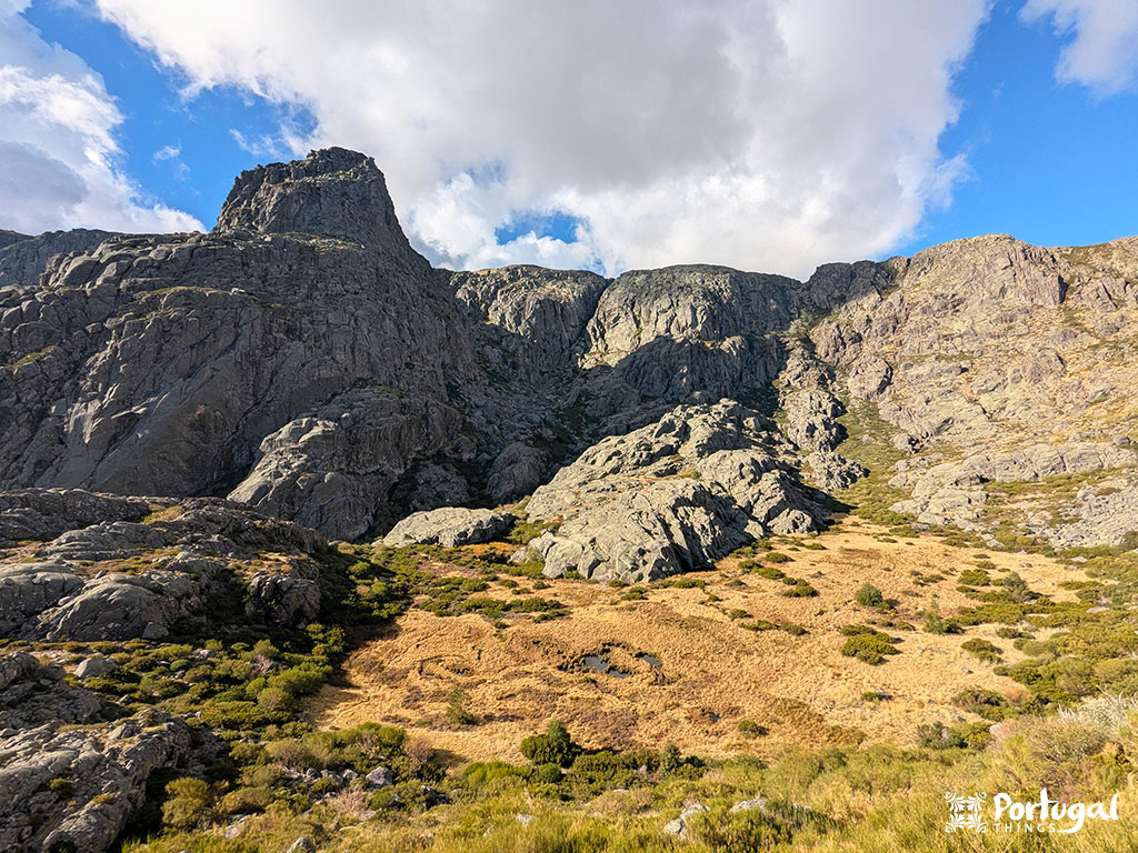 Penhascos de montanhas rochosas sob um céu parcialmente nublado com manchas de grama amarela e arbustos verdes em primeiro plano. Uma etiqueta 