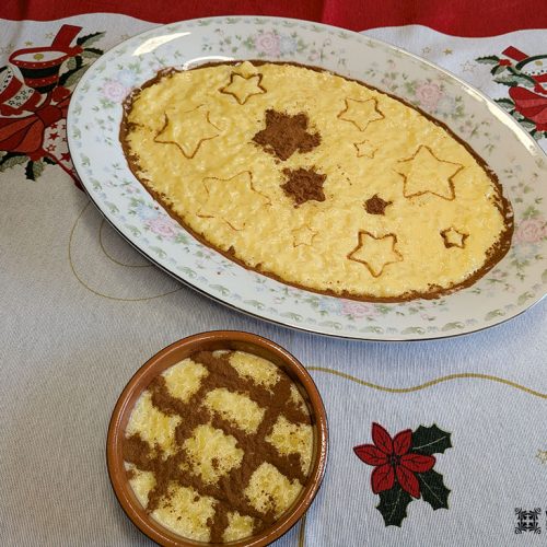 Uma travessa oval grande e uma tigela redonda pequena contêm Arroz-Doce, uma receita tradicional de pudim de arroz amarelo decorado com padrões de estrelas e grades feitos de canela, colocados em uma toalha de mesa com bordados com tema natalino.
