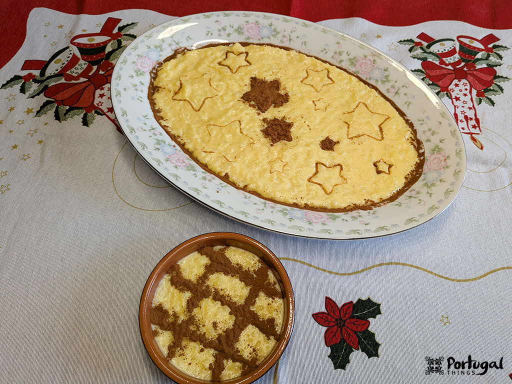A large oval dish and a small round bowl contain Arroz-Doce, a traditional yellow rice pudding decorated with cinnamon star and grid patterns, placed on a Christmas-themed embroidered tablecloth.