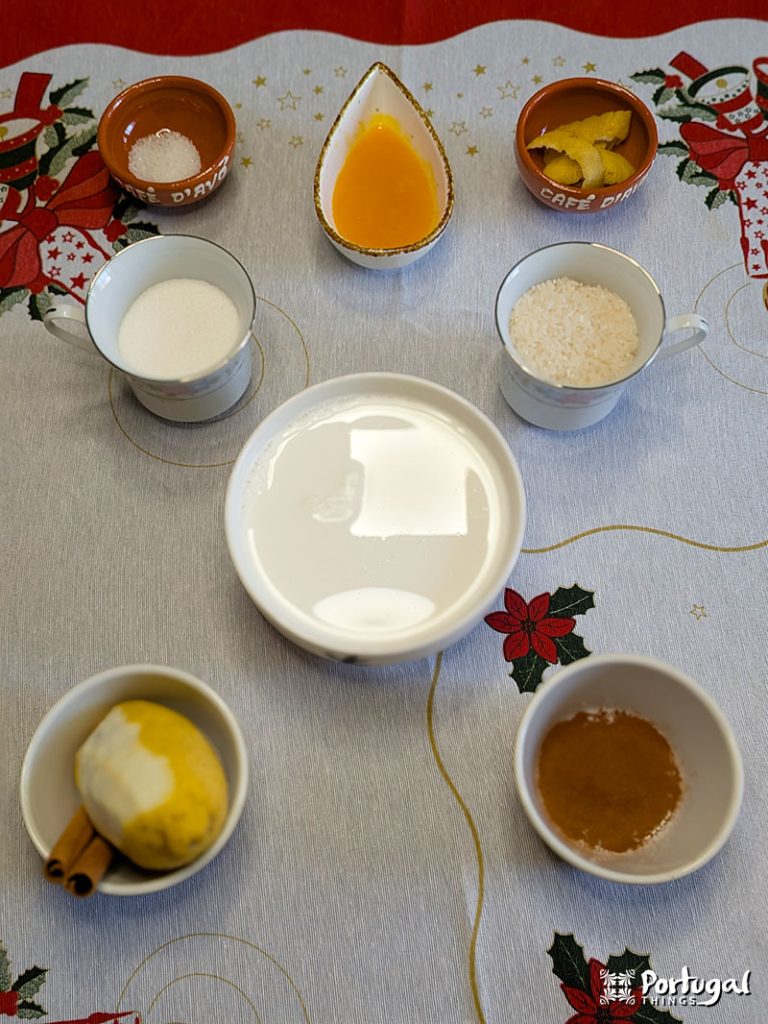 Several small bowls and cups containing ingredients for Arroz-Doce, including sugar, milk, rice, lemon, cinnamon sticks, ground cinnamon, salt, orange juice, and lemon zest, are arranged on a table with a festive tablecloth.