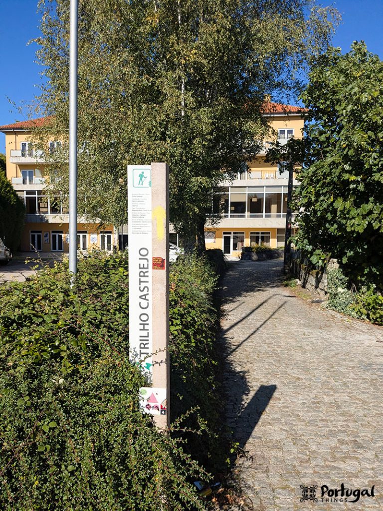 A stone path surrounded by green bushes leads to buildings with balconies in Castro Laboreiro. In the foreground, a vertical sign reading 