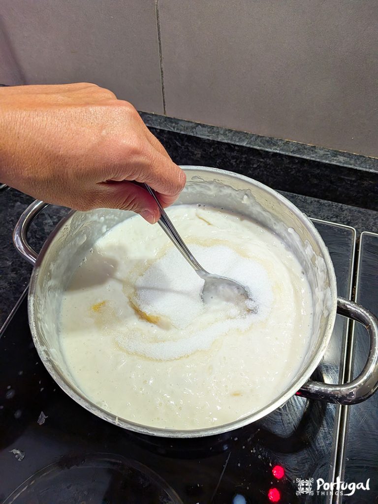 A hand stirs a pot of creamy white mixture with a spoon on a stovetop, stirring in granulated sugar. This classic Arroz-Doce, or rice pudding, simmers gently on a black electric stovetop—an irresistible traditional recipe.