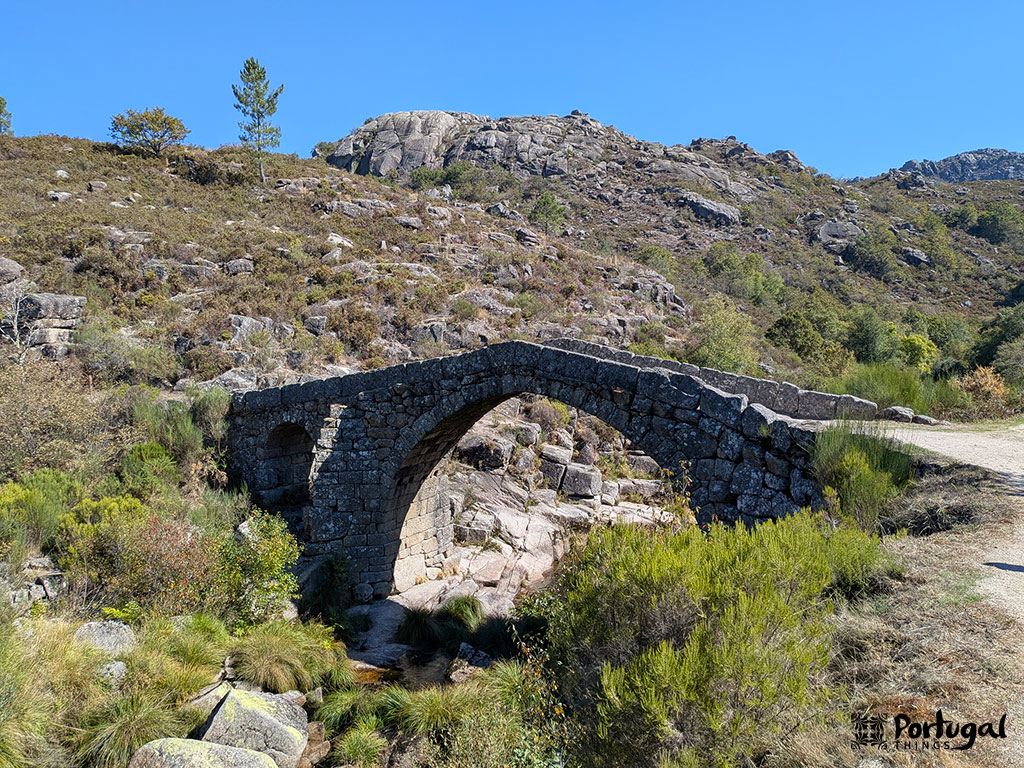 A small, ancient stone arch bridge crosses a rocky stream on the Trilho Castrejo Trail (PR3), surrounded by shrubs and hills under a clear blue sky. Rocky terrain and sparse trees appear in the background.