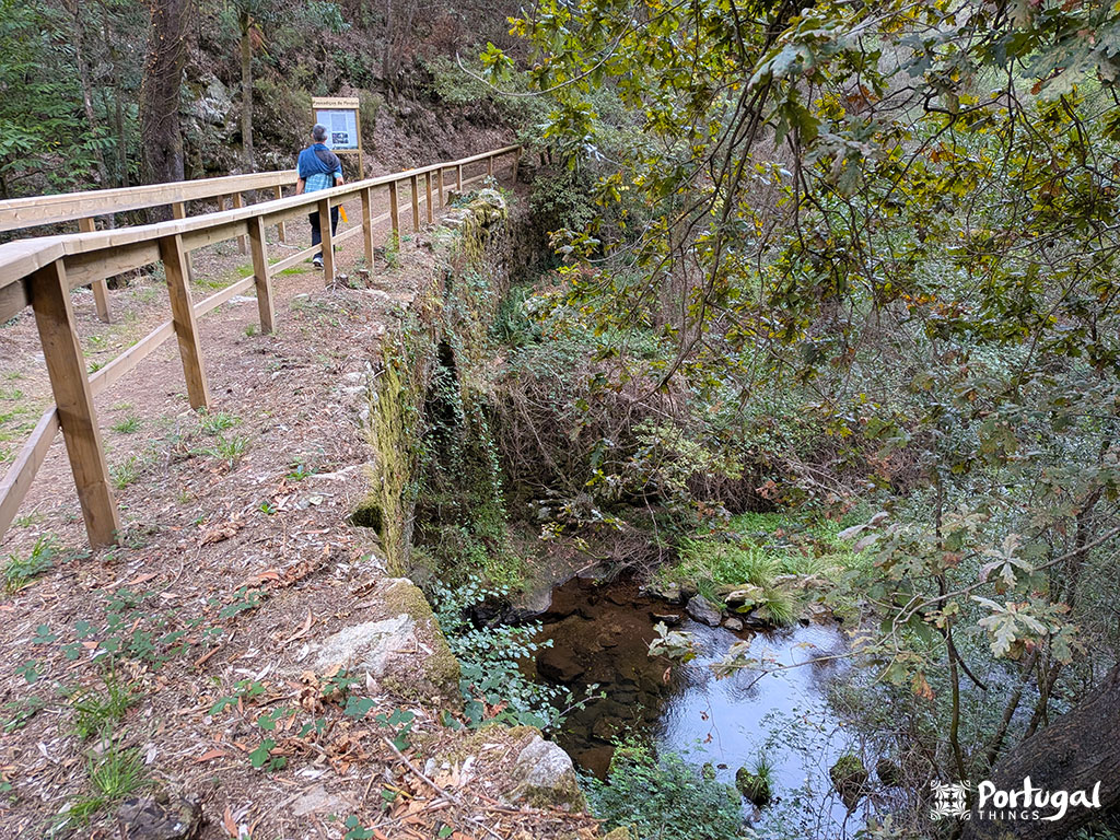 A wooden walkway, part of the Pindelo Walkways, runs alongside a stone wall above a small stream in a forested area. Two people walk along the trail, surrounded by lush trees and vegetation, with a sign visible ahead.