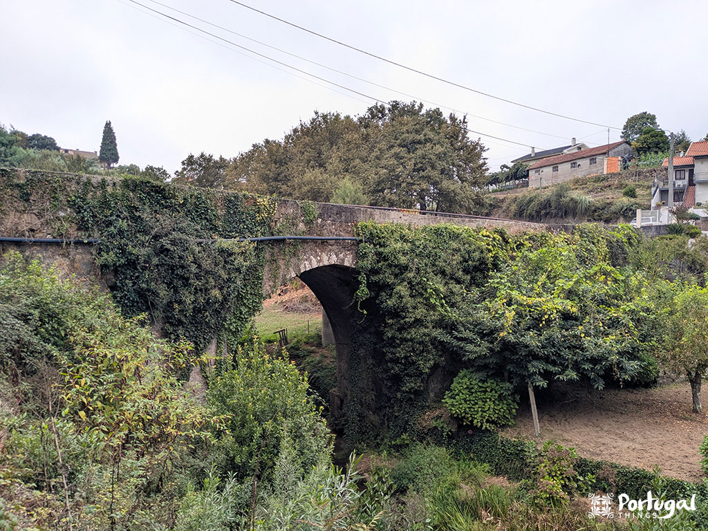 A stone bridge covered in ivy and vegetation crosses a small ravine near the Pindelo Walkways, surrounded by dense greenery. Houses lie to the right, while a cloudy sky approaches.