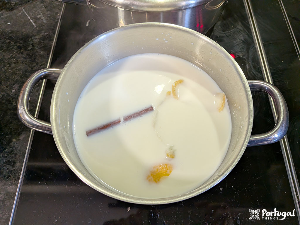 A metal pot on the stovetop contains milk, a cinnamon stick, lemon peel, and orange peel, boiling together—a classic start to Arroz-Doce, a traditional recipe. Steam rises, and a thin film of milk floats on the surface.