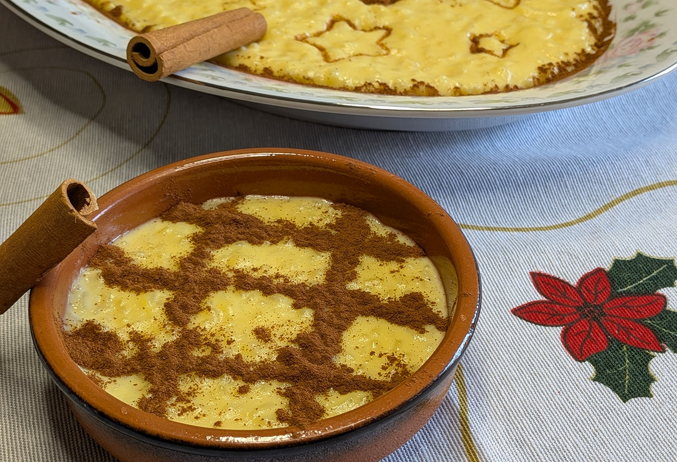 Uma tigela e um prato de Arroz-Doce, uma receita tradicional, coberta com canela, estão sobre uma toalha de mesa adornada com azevinho e poinsétia. Os paus de canela acompanham as sobremesas clássicas.